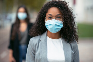 Two pretty business women wearing a hygienic face mask while looking at camera in the street....