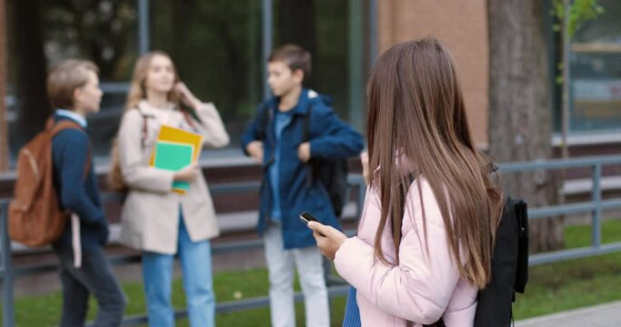 Portrait of happy schoolgirl walking on street to school and typing on cellphone. Female pupil waving hand to her classmates outdoors. Student texting and tapping on smartphone. School concept
