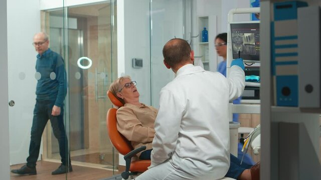 Stomatologist pointing on digital screen explaining x-ray to elderly woman. Doctor and nurse working together in modern stomatological clinic, examining, showing radiography of teeth on monitor