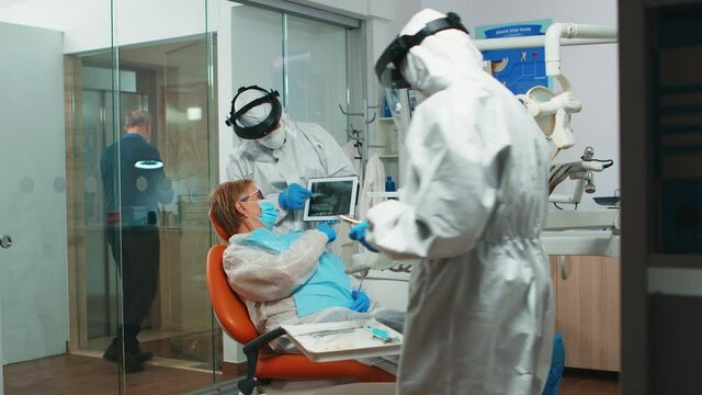 Dentist in protective equipment showing on tablet dental x-ray reviewing it with senior patient. Medical team wearing face shield coverall, mask, gloves, explaining radiography using notebook display
