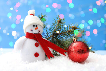 Cute snowman, Christmas ball and fir tree branch on snow against light blue background, bokeh effect
