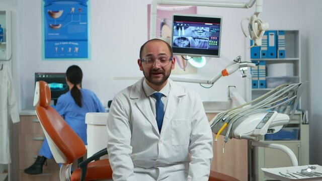 Man stomatologist speaking on video camera sitting on chair in dental clinic with assistent in background. Dentistry doctor looking on webcam explaining treatment while nurse is working on computer
