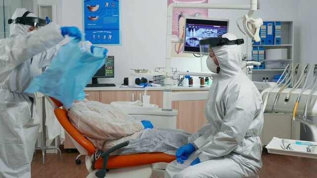 Dentist nurse in coverall putting dental bib to old woman before stomatological examination during covid-19 pandemic. Assistant and orthodontic doctor wearing protection suit, face shield mask gloves