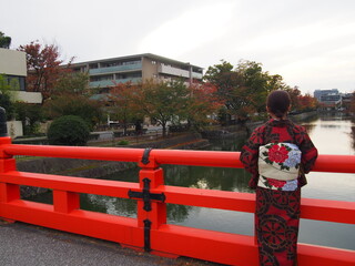 An Asian woman in a traditional Japanese kimono standing on a red bridge looking out at the river, Kyoto, Japan