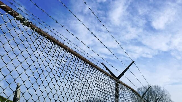 Chain-link fence with barbed wire, floating clouds in blue sky, time-lapse
