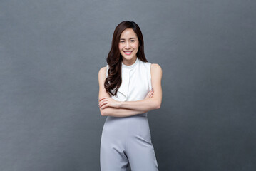 Portrait of smiling young beautiful Asian businesswoman looking at camera and doing arm crossed gesture in isolated studio gray background