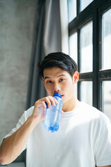 Fototapeta na wymiar Young man in white shirt drinking from bottle of water at home.