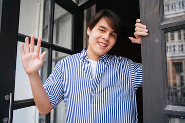 Asian man waving hand at camera at front door.