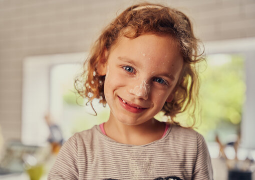 Portrait Of Cheerful Little Girl Cooking In Kitchen With Flour Applied On Face