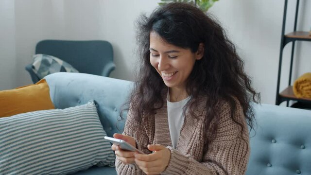 Hapy mixed race girl is using smartphone touching screen then smiling getting good news showing thumbs-up hand gesture sitting on sofa at home