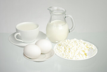 Milk in a jug and in a white cup, eggs, cottage cheese on a saucer on a gray background.