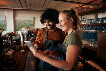 Cheerful male and female waiter wearing apron using digital tablet standing behind counter in cafe