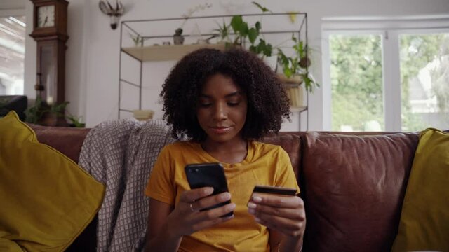 Zoom in shot of an african young woman sitting on sofa with crossed legs making online payment using credit card and smart phone