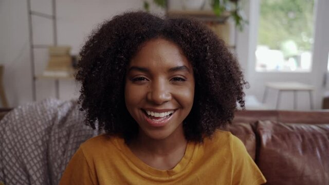 Friendly african millennial single woman sitting on sofa in living room smiling and looking at camera