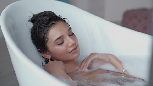 Close up relaxed woman touching skin with foam in bath. Top view of sexy girl taking bathtub at home. Romantic brunette woman relaxing bath in slow motion.