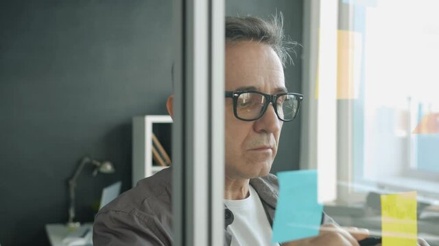 Mature guy is writing on bright sticky notes on glass board in office doing creative work for business development. People and workplace concept.