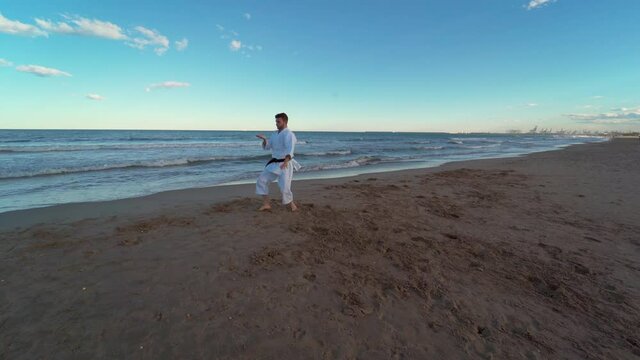 Karate fighter alternating fast and powerful and slow and fluid movements at beach with the waves in the background. Karate fighter kicking and punching. Karate fighter doing katas on the beach