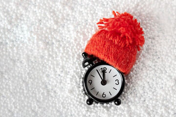 Black alarm clock in a red knitted hat on a white snow background. Concept of New Year year and Christmas
