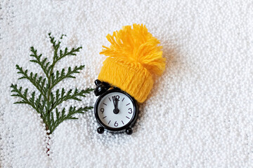 A black alarm clock in a yellow knitted hat and a spruce branch on a white snow background. The concept of New year and Christmas