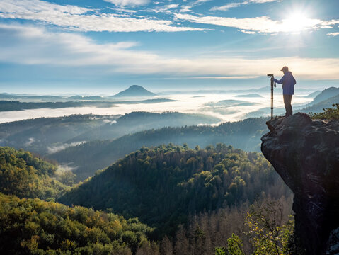 Photographer in silhouette carrying tripod with big camera on locky view above landscape