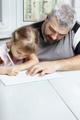 adult gray-haired father teaches little daughter to write at the table at home. family values concept