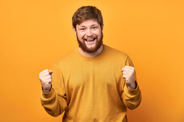 Pleased young caucasian bearded man rejoices to hold his hands up celebrating being a winner,...