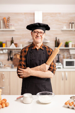 Caucasian old man wearing apron in home kitchen smiling at camera. Retired elderly baker in kitchen uniform preparing pastry ingredients on wooden table ready to cook homemade tasty bread, cakes and