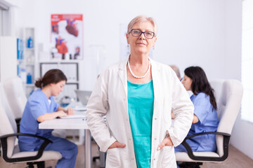 Portrait of senior doctor smiling at camera in hospital conference room with medical staff in the...