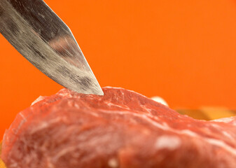 Close-up shot of a butcher's knife and a piece of red raw pork meat on a bright orange background....