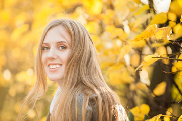 Portrait of a beautiful young woman against a background of colorful golden foliage in an autumn park. Attractive woman with light brown hair near bushes with yellow leaves. Fall season. Close-up.