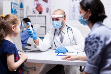 Fototapeta premium Doctor wearing face mask against coronavirus checking temperature using digital thermometer on child during medical examination. Health pediatrician specialist providing health care services.