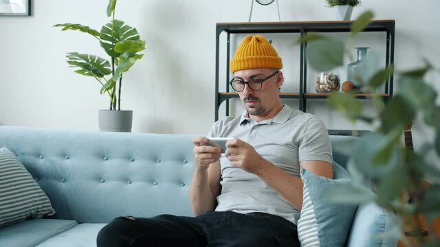 Young hipster is playing video game losing competition throwing smartphone feeling mad sitting on couch at home. Gaming and negative emotions concept.