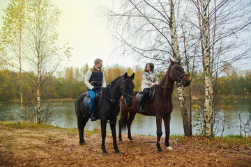 Cute young couple on horsebacks in the autumn forest by lake. Riders in autumn Park in inclement cloudy weather with light rain. Concept of outdoor riding, sports and recreation. Copy space