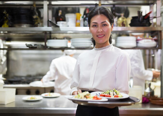 positive female waitstaff standing with tray in kitchen in restaurante