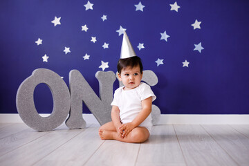 One year old baby boy celebrates birthday near silver letters ONE on blue background with stars