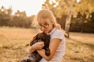 Teenager blonde girl with big glasses laughing and playing with little puppy spaniel in the warm park
