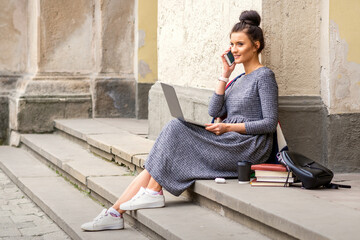 Young adult female student talking by smartphone sitting on stairs with laptop near university building