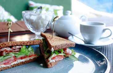 sandwiches with bacon herbs and tomatoes on a dark plate with a white Cup of tea a teapot and a sugar bowl in the background with a blurred background