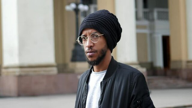 Urban portrait of mixed race man with glasses, standing on the street dressed traditional hat and looking at the camera in slow motion