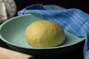 Proofing process of homemade bread dough on enamel ware, cover with blue napkin, with rolling pin and baking timer on wooden dark background. 