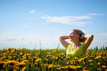 Fototapeta premium Pretty woman on a field with green grass and yellow dandelion flowers in a sunny day. Girl on nature with yellow flowers and blue sky.