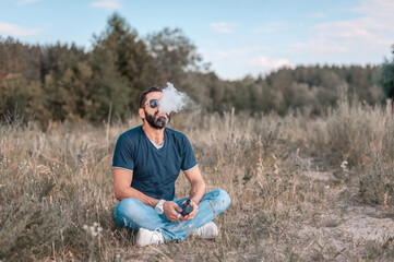 Naklejka na ściany i meble Bearded man is resting in a forest clearing and vaping and letting off steam from an vape. Vaping activity.