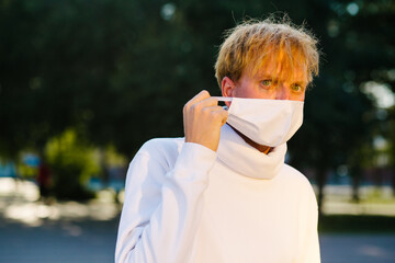 A young man in a medical protective mask, anxiously looking to the side, adjusting the mask.