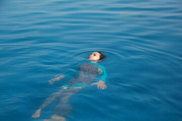 Joyful boy swimming with backstroke style on the infinity pool and sea view on the background