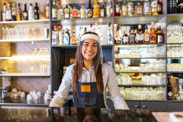 Portrait of a beautiful bartender standing at the counter smiling and looking at the camera while while wearing face shield due to covid-19, shelves full of bottles with alcohol on the background