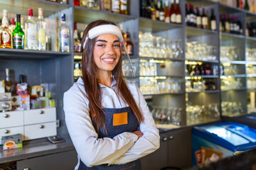 Portrait of a beautiful bartender standing at the counter smiling and looking at the camera while while wearing face shield due to covid-19, shelves full of bottles with alcohol on the background