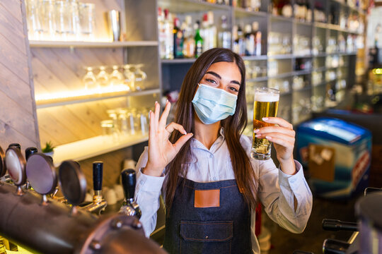Bartender with protective face mask,covid-19 protection, serving a draft beer at the bar counter during coronavirus pandemic, showing OK sign,shelves full of bottles with alcohol on the background