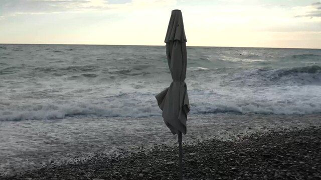 Storm at the sea coast. Closed beach umbrella, strong wind, big waves with lot of foam. Summer evening, bright dky and gray water.