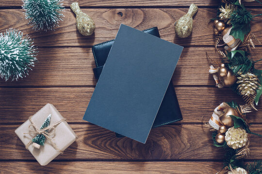 Mockup blank black book cover with Christmas gift box, Xmas ornaments and Christmas tree model decor on wooden table background. Flat lay, Top view with copy space