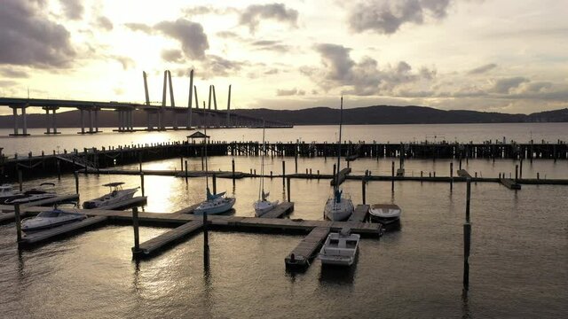 An aerial shot of the Tarrytown Marina with the Mario M. Cuomo Bridge in the background. The drone camera dolly in over the marina & river towards the bridge during a golden sunset with clouds.